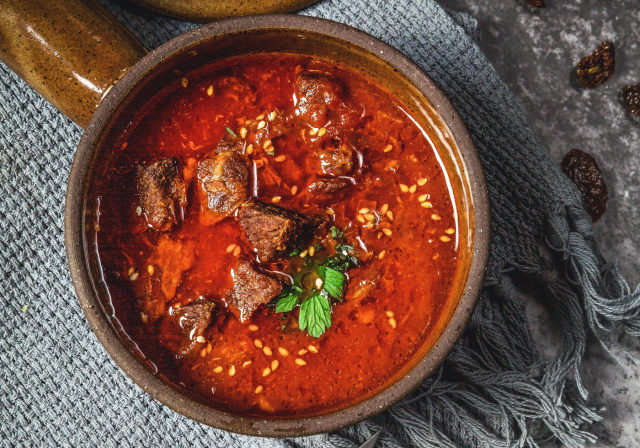 Kräftig rote Suppe mit Rindfleischwürfeln in einer Steingutschale, mit frischen Kräutern und Sesam bestreut
