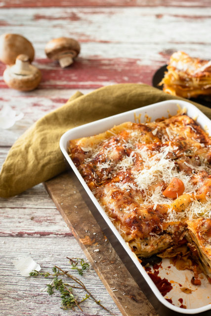 Pilzlasagne in einer weißen Auflaufform, mit Käse bestreut, daneben Champignons